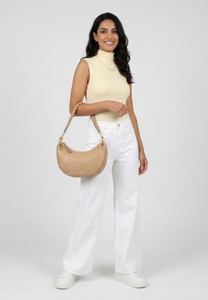 Femme aux longs cheveux foncés portant un col roulé beige sans manches, un pantalon blanc à jambes larges, des baskets blanches, et tenant un sac à main beige à poignée tressée.