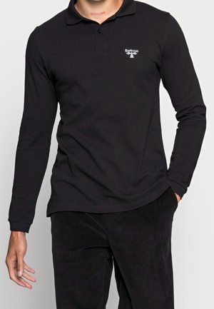 Mand iført en sort langærmet polo med Barbour-logo på brystet og sorte bukser, med den ene hånd i lommen.