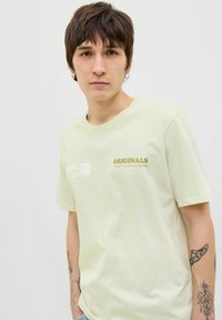 Lichtgroene katoenen t-shirt met korte mouwen, ronde hals en tekst op de voorkant: "ORIGINALS" in gouden letters. Textuurstof.