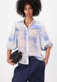 Blus ringan, transparan dengan pola bunga biru, dilengkapi dengan kancing di depan dan lengan puff. Dipadukan dengan celana navy bermotif dan dompet hitam.