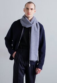 Pilkas vilnonis šalikas apvyniotas aplink kaklą, derinamas su tamsiai mėlynu tekstūruotu kardiganu ir pritaikytomis juodomis kelnėmis. Paprastas dizainas, minkšta tekstūra.