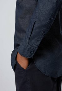 Mörkblå button-up skjorta med en strukturerad tyg, rullad ärmdetalj och en böjd fåll. Bärs över mörka byxor.