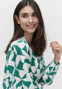 Grön och vit mönstrad blus med en flödande design, med rund halsringning och långa ärmar. Tyget verkar mjukt och lätt.