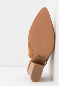 Chaussure à talon haut en suède marron clair avec un bout pointu et une semelle texturée. Présente un talon incurvé et un design minimaliste.