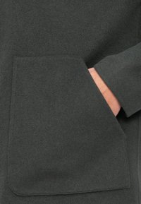 Mørkegrå, strukturert ullfrakk med en stor, lapplomme foran og en hånd delvis synlig inne i lommen.