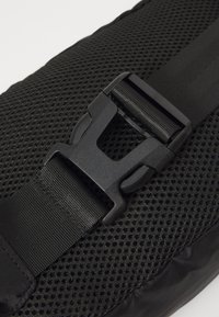 Gros plan sur un sac à dos noir présentant une surface en maille texturée et une boucle en plastique noir, avec un matériau de sangle lisse et des accents subtils.