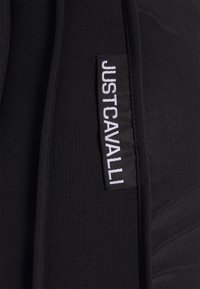 Černá látková panel s vertikálním bílým logem "JUSTCAVALLI". Materiál má hladkou strukturu s jemným leskem.