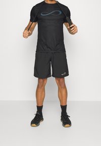 Sort atletisk t-shirt med blå uendelighedslogo, parret med sorte shorts og sorte sneakers med en tan sål. Begge dele er lavet af letvægtsstof.