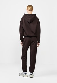 Asmuo su šviesiais plaukais surišusiais į kuodą, vilkintis tamsiai rudą kapsulę ir atitinkamus sportinius džinsus, stovi atsisukęs nugara prieš neutralų foną.