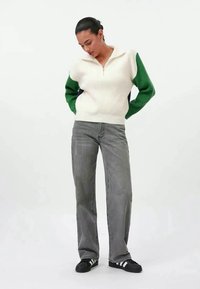 Gestrickter Pullover mit halbhohem Reißverschluss in Cremefarben, mit grünen Ärmeln. Kombiniert mit grauen Jeans mit geradem Bein und schwarzen Sneakers mit weißen Streifen.