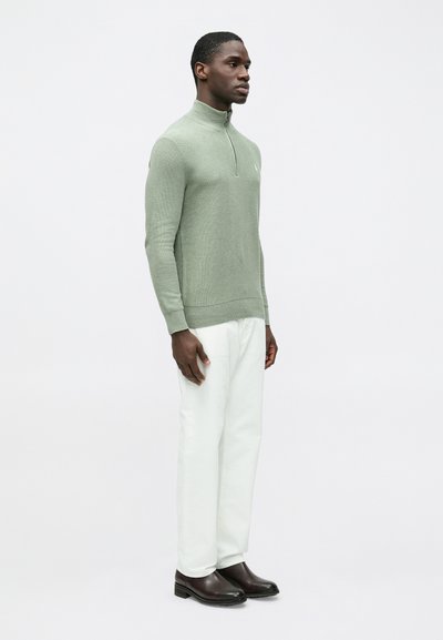 Világoszöld kötött pulóver félcipzáras gallérral és bordázott szegéllyel, fehér nadrággal és sötétbarna csizmával párosítva.