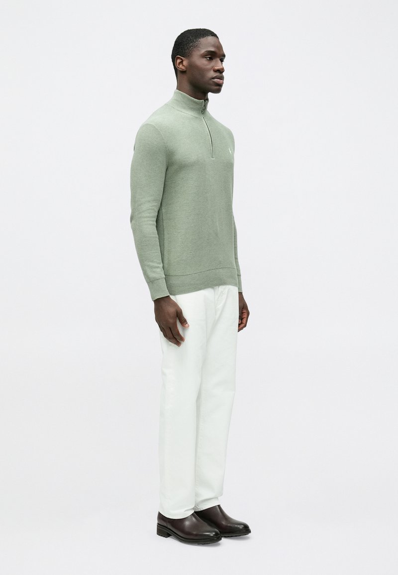 Maglione verde chiaro lavorato a maglia con collo mezza zip e orlo a coste, abbinato a pantaloni bianchi e stivali marrone scuro.