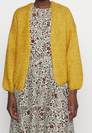 Cardigan jaune tricoté avec un fil texturé, à front ouvert et manches bouffantes. Porté par-dessus une robe à motifs floraux dans des couleurs atténuées.