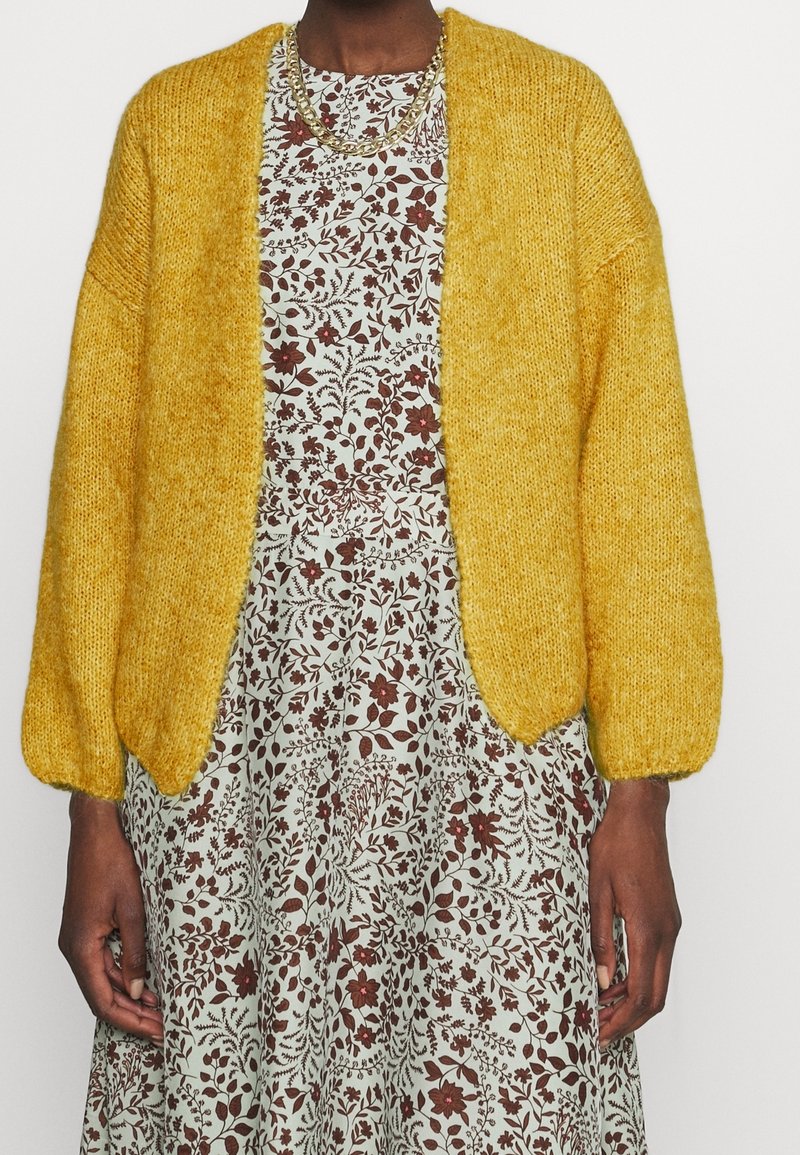 Cardigan jaune tricoté avec un fil texturé, à front ouvert et manches bouffantes. Porté par-dessus une robe à motifs floraux dans des couleurs atténuées.