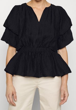 Femme portant un chemisier noir texturé avec des manches courtes bouffantes et une taille froncée, associé à un pantalon beige clair.