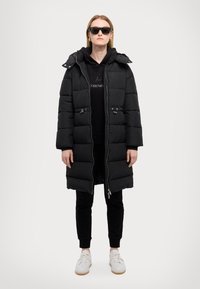 Piumino nero con zip frontale e cappuccio, caratterizzato da un design trapuntato, abbinato a una felpa nera, pantaloni neri e sneakers bianche.