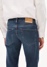 Jeans de mezclilla en azul oscuro con una textura suave, que presentan una etiqueta marrón en la parte trasera, costuras en contraste y dos bolsillos traseros.