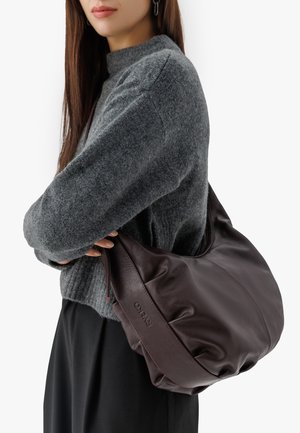 Bordowa skórzana torebka na ramię o miękkim, teksturowanym wykończeniu, z zamkiem błyskawicznym i delikatnym logo. Założona na szary sweter.
