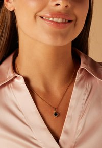 Collier en or avec un pendentif noir, featuring une bordure décorative. Le mannequin porte une chemise en satin rose clair, mettant en valeur les détails du collier.