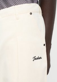 Un pantalon crème en tissu durable qui présente une poche latérale avec un bouton en métal noir et "Tacker" brodé en cursive.