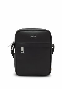 Svart syntetisk crossbody-väska med slät textur, silverfärgad logotyp, justerbar rem och ett dragkedjefack. Kompakt fyrkantig form.