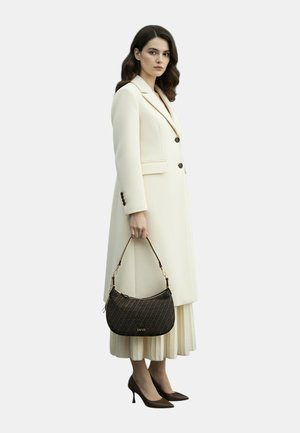 Donna con un lungo cappotto crema e una gonna plissettata che tiene una borsa scura con decorazioni, indossa tacchi marroni a punta, in piedi contro uno sfondo semplice.