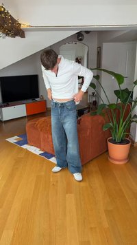 Asmuo dėvi baltą ilgomis rankovėmis marškiniu ir laisvas mėlynas džinsus, stovi ir tvarko juosmens juostą; fone matyti žalias augalas ir rudai spalvos sofa.