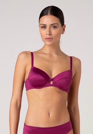 Donna con capelli scuri legati indietro che indossa un coordinato di reggiseno e biancheria intima in raso magenta su uno sfondo uniforme.