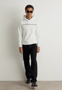 Hanorac alb cu glugă, având un logo și o linie negre, purtat cu pantaloni negri și pantofi negri. Modelul poartă ochelari de soare negri. Fundal simplu.