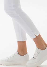 Vita cropped byxor med en knappeaccent vid fållen, kombinerat med vita sneakers. Slät tygtextur och avsmalnande bendesign.