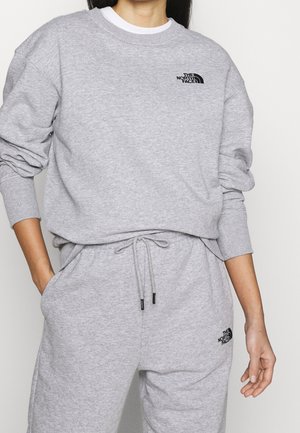 Femme portant un sweat-shirt et un pantalon de survêtement assortis gris clair The North Face avec un logo noir sur la poitrine et la cuisse, debout avec une main dans la poche.