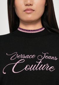 Črn pleten pulover z rožnato vezenim napisom "Versace Jeans Couture" in dekorativnim ovratnikom iz perlic v odtenkih roza in vijolične.