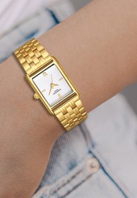 Kuldne ristkülikuline käekell, millel on valge tekstuuriga kellanäidik, kuldsed tundmärkmed ja lülitustega käevõru. Kannab päevitunud randmel, denimist tausta ees.