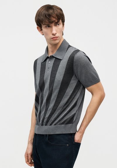 Νεαρός άνδρας με κοντά καστανά μαλλιά, φορούν γκρι πόλο μπλούζα με σκούρες κάθετες ρίγες και σκούρο παντελόνι, στέκεται με το ένα χέρι στην τσέπη.