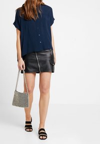 Blusa a maniche corte blu navy, abbinata a una mini gonna di pelle nera con zip frontale. Accessoriata con una borsa a fantasia e sandali neri.