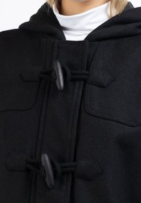 Czarny wełniany płaszcz z kapturem, z zapięciem na toggle i dwoma przednimi kieszeniami. Gładka tekstura z detalami ściegów na brzegach.