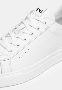 Zapatilla blanca con piel sintética suave, diseño de cordones, detalles de perforación en el costado y un logotipo negro en la lengüeta.