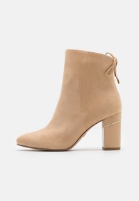 Bottine beige en daim avec un bout pointu, un talon bloc mi-hauteur orné d'un détail doré, et un lien à l'arrière pour faciliter l'enfilage.