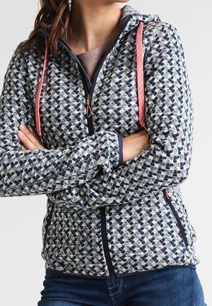 Kobieta ubrana w niebiesko-białą, kraciastą bluzę z kapturem na zamek, z koralowymi sznurkami, skrzyżowanymi ramionami i niebieskich dżinsach na prostym tle.