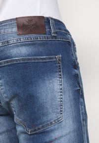 Blue denim jeans featuring a brown leather patch at the waist with embossed logo, subtle distressing, and a rear pocket accent.