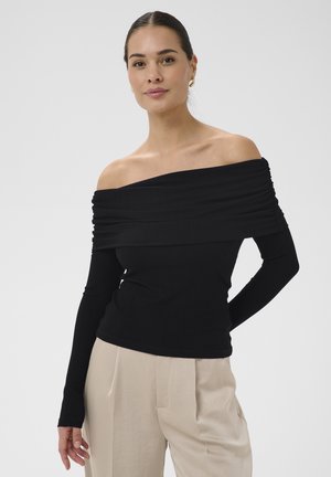 Ženska s temnimi lasmi, oblečena v črno rebrasto majico brez ramen in bež hlače, stoji z eno roko za hrbtom ob enobarvnem ozadju.