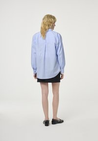 Light blue, long-sleeve, button-up shirt with a relaxed fit, rounded hem, and back pleat, paired with a black mini skirt and flat shoes.