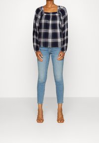 Blouse à carreaux noir et blanc avec décolleté carré et manches bouffantes, associée à un jean bleu slim et des sandales à brides de couleur tan.