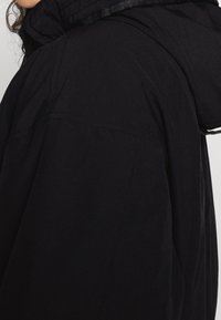 Svart huva jacka med en avslappnad passform, synliga sömmar och en mjuk textur. Huvan och kragen har kontrasterande kantdetaljer.