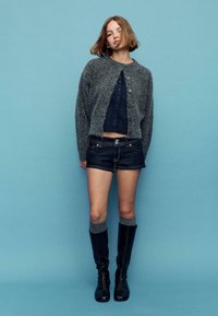 Sivi pleteni kardigan prek mornarsko modernega kratkega topa, kombiniran s temnimi denim kratkimi hlačami in črnimi škornji do kolen s sivimi nogavicami. Monotona modra podlaga.