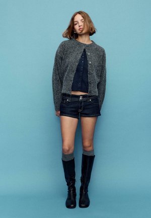 Cardigan en tricot gris porté sur un haut court bleu marine, associé à un short en denim foncé et des bottes noires montantes avec des chaussettes grises. Fond bleu uni.