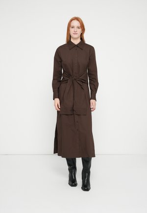 Καφέ πουκάμισο-φόρεμα με κουμπιά, μακριά μανίκια και ζώνη στη μέση· διαθέτει ίσια γραμμή, σκίσιμο στο πλάι και μαύρα μποτάκια σε αντίθεση.