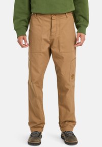 Tan work pants with a straight fit, durable fabric, multiple pockets, and an embroidered logo. Paired with a green sweatshirt and boots.