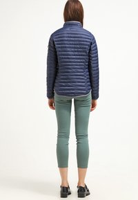 Giacca a vento blu navy con cuciture orizzontali, collo alto e una texture liscia e lucida. Indossata sopra pantaloni aderenti verde chiaro e scarpe nere.