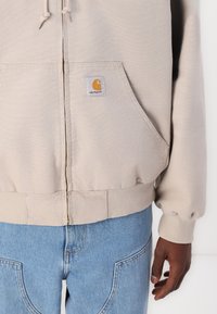 Beige zip-up hoodie met ribbelboorden en -onderkant, voorzien van twee voorzakken en een vierkant Carhartt-logo in geel en wit. Draag het met blauwe jeans.